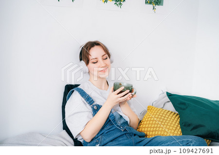 Woman with closed eyes listen music and enjoy cup of coffee or tea. Calm female spend free time at home enjoy favourite song with wireless modern headphones. Pastime weekend relax, no stress concept Woman with closed eyes listen music and enjoy cup of coffee or tea. Calm female spend free time at home enjoy favourite song with wireless modern headphones. Pastime weekend relax, no stress concept 109427691