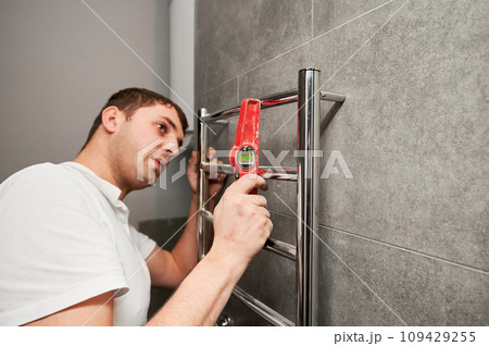 Male plumber checking electric heated towel rack with spirit level ruler in bathroom. Man installing stainless steel bath towel heater in apartment under renovation. Male plumber checking electric heated towel rack with spirit level ruler in bathroom. Man installing stainless steel bath towel heater in apartment under renovation. 109429255