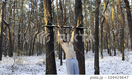 Strong and muscular man doing exercises on horizontal bar at winter forest. Young sportsman muscling up at beautiful nature. Athletic guy training outdoor. Concept of sport and active lifestyle Strong and muscular man doing exercises on horizontal bar at winter forest. Young sportsman muscling up at beautiful nature. Athletic guy training outdoor. Concept of sport and active lifestyle 109429647