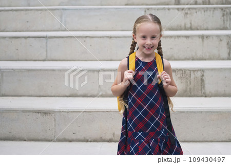 In her school dress and yellow backpack, a happy blonde schoolgirl smiles with pure joy as she embarks on her journey to school 109430497
