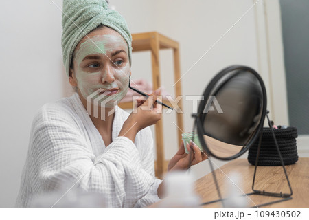 Facial Bliss: She, in a bathrobe, applies a clay mask with a brush, gazing at the mirror, indulging in her organic facial treatment at home. Facial Bliss: She, in a bathrobe, applies a clay mask with a brush, gazing at the mirror, indulging in her organic facial treatment at home. 109430502