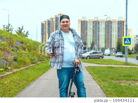 A cute man stands with a skateboard and a phone in his hand and smiles at the camera on a city street. A man with a plaid shirt stopped to call on his skateboard. Concept of a man with a skateboard. A cute man stands with a skateboard and a phone in his hand and smiles at the camera on a city street. A man with a plaid shirt stopped to call on his skateboard. Concept of a man with a skateboard. 109434111