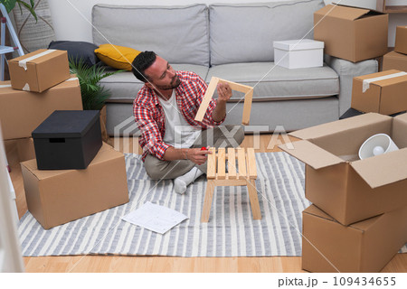 Brunette man with beard tries to figure out correct assembly of wooden chair sitting with serious expression on floor near sofa among cardboard boxes in spacious premise Brunette man with beard tries to figure out correct assembly of wooden chair sitting with serious expression on floor near sofa among cardboard boxes in spacious premise 109434655