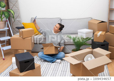 Woman takes selfies with box close to sofa in modern house waiting for lovely boyfriend from work to help sort out things needed to decorate contemporary cottage house Woman takes selfies with box close to sofa in modern house waiting for lovely boyfriend from work to help sort out things needed to decorate contemporary cottage house 109434657