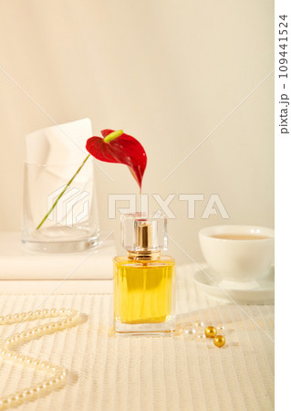Front view of a yellow perfume bottle displayed on beige background with vase of red flower, cup of coffee and pearl necklace. Scene for advertising essential oil product with mockup for design 109441524