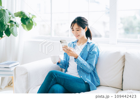 ソファに座ってコーヒーを飲みながらスマホを見る女性 ソファに座ってコーヒーを飲みながらスマホを見る女性 109442423
