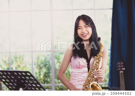 Young woman saxophonist with an instrument stands by the window. Cozy home interior. Concept of hobbies, inner world, development of talents, music school. Young woman saxophonist with an instrument stands by the window. Cozy home interior. Concept of hobbies, inner world, development of talents, music school. 109444502