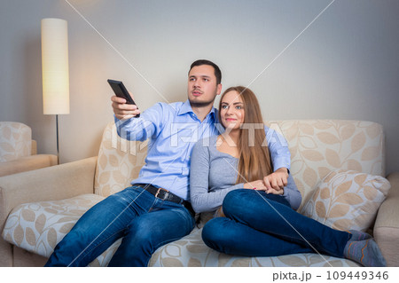 Happy couple sitting on sofa watching television together Happy couple sitting on sofa watching television together 109449346