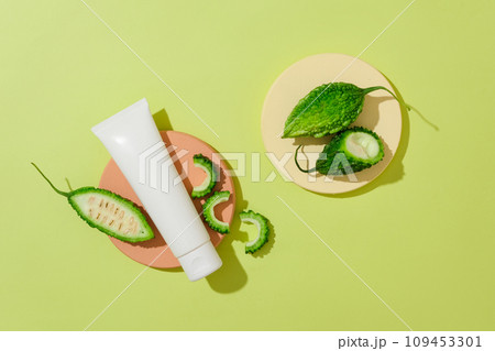Creative scene for advertising cosmetic with ingredient from bitter melon extract. A white plastic tube on orange podium and fresh bitter melon slices on beige podium decorated on green background 109453301