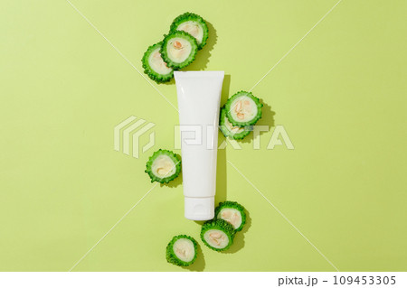 Top view of bitter slices decorated with cosmetic bottle unbranded on green background. Mockup for design and advertising cosmetic of bitter melon extract. Space for design 109453305