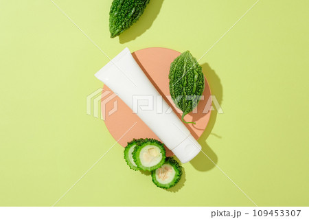 An unbranded plastic tube orange podium with fresh slices of bitter melon decorated on green background. Scene for cosmetic product of natural extract presentation. Space for design, top view 109453307