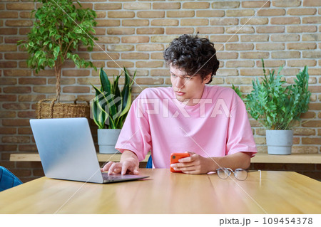 Young male college student sitting at desk at home with laptop and smartphone Young male college student sitting at desk at home with laptop and smartphone 109454378