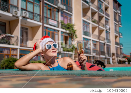Woman pool Santa hat. A happy woman in a blue bikini, a red and white Santa hat and sunglasses poses near the pool with a glass of champagne standing nearby. Christmas holidays concept. Woman pool Santa hat. A happy woman in a blue bikini, a red and white Santa hat and sunglasses poses near the pool with a glass of champagne standing nearby. Christmas holidays concept. 109459003