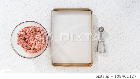 Flat lay. Scooping ground meat with dough scoop into a baking sheet lined with parchment paper to prepare oven-baked meatballs. Flat lay. Scooping ground meat with dough scoop into a baking sheet lined with parchment paper to prepare oven-baked meatballs. 109461127