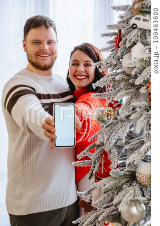 man with woman holding phone with empty screen standing near christmas tree 109463600