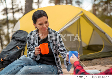 Beautiful adult girls drinking tea in camping in forest with tent on background and smiling. Young woman traveller during hiking in mountains resting Beautiful adult girls drinking tea in camping in forest with tent on background and smiling. Young woman traveller during hiking in mountains resting 109463827