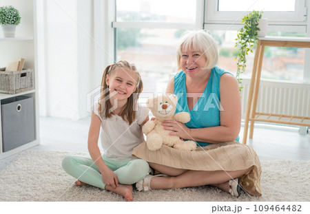Cute little girl playing plush teddy bear toy with mature woman sitting on floor in modern apartment 109464482