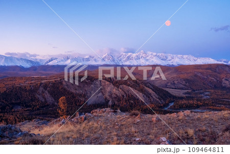 Dawn in the mountains with a full moon in the sky. Impressive panorama of dawn snow-capped peaks and a wide mountain valley with winding river in a canyon in soft purple tones. Dawn in the mountains with a full moon in the sky. Impressive panorama of dawn snow-capped peaks and a wide mountain valley with winding river in a canyon in soft purple tones. 109464811