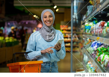 Satisfied Arab woman in hijab is shopping in the store, holding a phone and a shopping basket, smiling and looking at the camera. Satisfied Arab woman in hijab is shopping in the store, holding a phone and a shopping basket, smiling and looking at the camera. 109467482