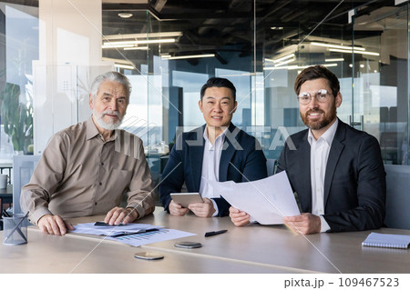 Three adult businessmen in suits sitting at the table in the office, having a work meeting, smiling and looking at the camera. Three adult businessmen in suits sitting at the table in the office, having a work meeting, smiling and looking at the camera. 109467523