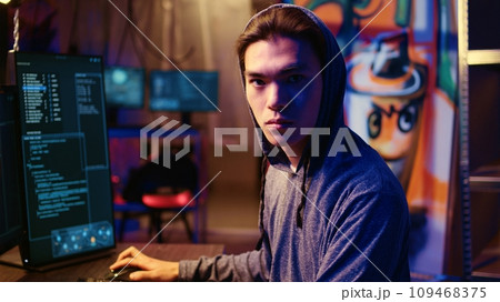 Portrait of asian hacker running code in graffiti painted basement, looking to find exploits for system vulnerabilities to be used against system owner for ransom, zoom in close up shot Portrait of asian hacker running code in graffiti painted basement, looking to find exploits for system vulnerabilities to be used against system owner for ransom, zoom in close up shot 109468375