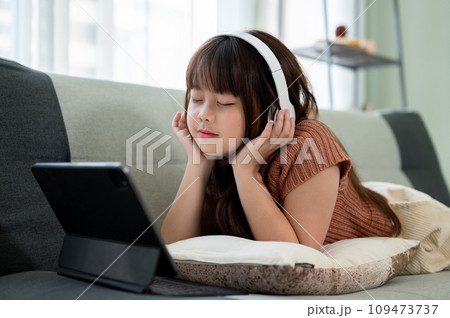 Young pretty girl is lying on a sofa and enjoying listening to music on her headphones. Young pretty girl is lying on a sofa and enjoying listening to music on her headphones. 109473737
