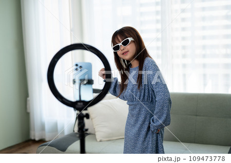 A cheerful young Asian girl in a cute dress and sunnies is recording her dance video at home. A cheerful young Asian girl in a cute dress and sunnies is recording her dance video at home. 109473778