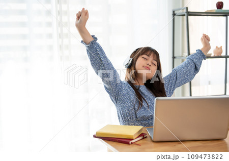 A cute young Asian girl is feeling tired, stretching her arms while studying online at home. A cute young Asian girl is feeling tired, stretching her arms while studying online at home. 109473822