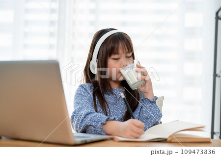 An adorable young Asian girl is drinking a glass of milk while doing homework at a table in her room An adorable young Asian girl is drinking a glass of milk while doing homework at a table in her room 109473836