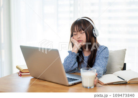 A cute young Asian girl is falling asleep while studying online at a table in her room. A cute young Asian girl is falling asleep while studying online at a table in her room. 109473840