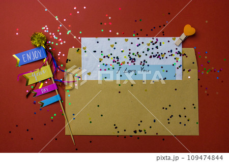 Love letter. A brown envelope with a Valentine's Day declaration of love note inside lies on a red terracotta background. Scattered colorful confetti. Festive greeting card for February 14 top view. 109474844