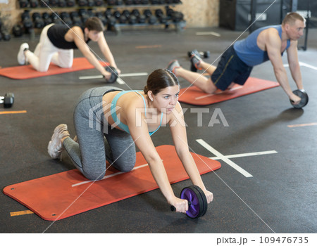 Motivated young woman practicing exercises with abs roller near other people in gym Motivated young woman practicing exercises with abs roller near other people in gym 109476735