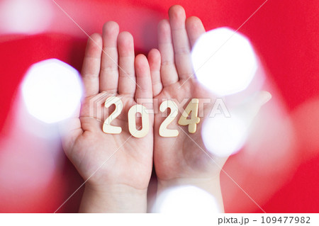 Child hands holding wooden number 2024 on palms on festive red background with copy space 109477982