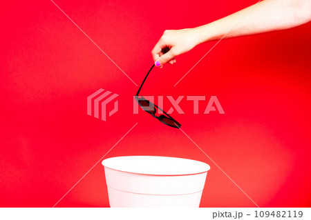 Hand throws sunglasses into trash bin with package on red background 109482119
