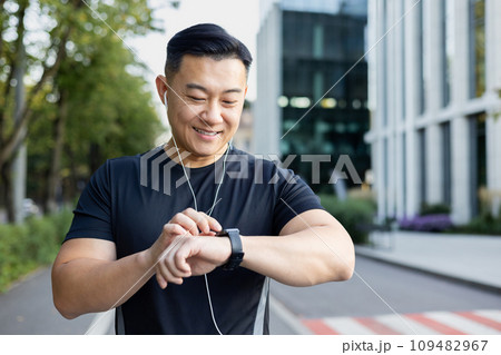 Close-up photo. Young Asian male athlete standing on city street wearing headphones and looking at watch. 109482967