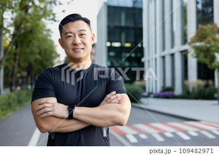 Portrait of a smiling young Asian male athlete standing on a city street, looking at the camera with his arms crossed. 109482971