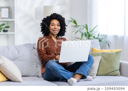 Portrait of a young African American female student sitting at home on the sofa in the lotus position and studying by correspondence on a laptop, looking at the camera with a smile. 109483094