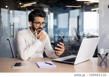 Pensive young Indian man businessman sits in the office at the table, looks at the phone, reads messages, solves questions. Pensive young Indian man businessman sits in the office at the table, looks at the phone, reads messages, solves questions. 109483116