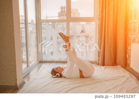 Side view portrait of relaxed woman listening to music with headphones lying on carpet at home. She is dressed in a white tracksuit. 109483297