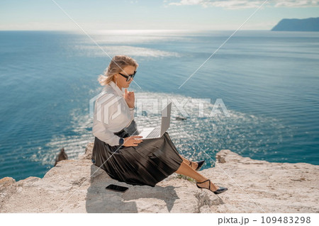 Business woman on nature in white shirt and black skirt. She works with an iPad in the open air with a beautiful view of the sea. The concept of remote work. Business woman on nature in white shirt and black skirt. She works with an iPad in the open air with a beautiful view of the sea. The concept of remote work. 109483298