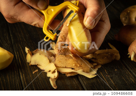 Close-up of a chef hands using a potato peeler to peel raw potatoes to prepare lunch. Vegetable menu concept for restaurant or hotel on dark background 109486608