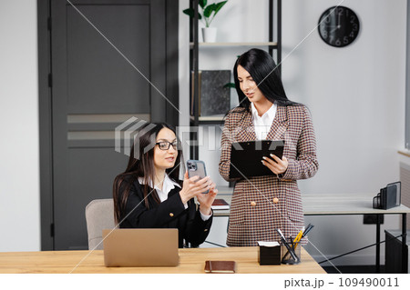 Two young attractive women are working in the office. A woman is talking on the phone, and her colleague is making notes in a notebook 109490011