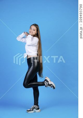 Happy woman with very long healthy hair posing, while flirty bending one leg. Side view of cheerful slim lady in leggings holding hands under chin and smiling, isolated on blue. Concept of posing. Happy woman with very long healthy hair posing, while flirty bending one leg. Side view of cheerful slim lady in leggings holding hands under chin and smiling, isolated on blue. Concept of posing. 109493790