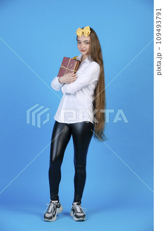 Pretty teen girl in yellow polka dot bow smiling, while hugging bunch of old books. Front view of nice female student with long hair posing, while carrying books, isolated on blue. Concept of posing. Pretty teen girl in yellow polka dot bow smiling, while hugging bunch of old books. Front view of nice female student with long hair posing, while carrying books, isolated on blue. Concept of posing. 109493791