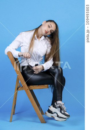 Pensive caucasian female with long fair hair wearing white shirt and leggings, holding glasses, tilting head to side, while looking at camera, isolated on blue studio background. Concept of posing. 109493803