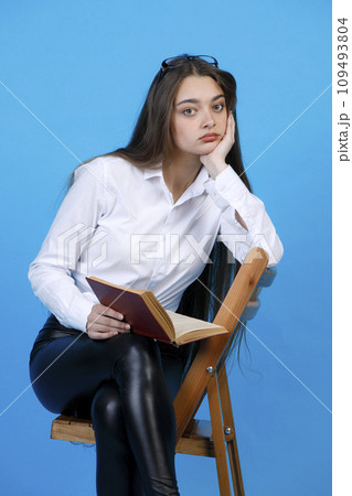Bored caucasian woman with glasses on head, resting head on hand, while sitting on chair indoor. Portrait of upset girl with open book, while looking at camera, isolated on blue. Concept of emotions. 109493804