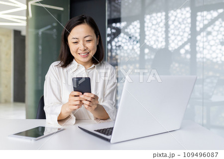 Young beautiful woman at workplace holds phone in her hands, Asian business woman reads online e-mail, female worker with a laptop inside the office browses the Internet pages, smiles contentedly. 109496087