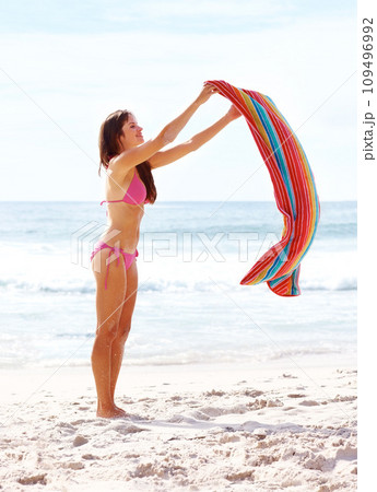 Woman, happy and towel on sand at beach for relax, sun tan and sunshine with bikini outdoor in summer. Person, smile and swimsuit by ocean with swimming, peace and wellness on holiday or vacation 109496992
