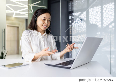 Successful smiling businesswoman working inside office at workplace, using laptop for video call, smiling. An Asian woman uses a headset to consult and communicate with a client. 109497393