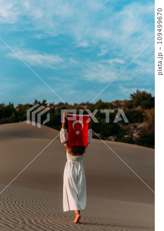 A beautiful dark-haired woman holds a Turkish flag in her hands, rear view. A woman is traveling in Turkey. A tourist on the background of Patara beach in Turkey. Vacation in Turkey. Turkish flag 109499670